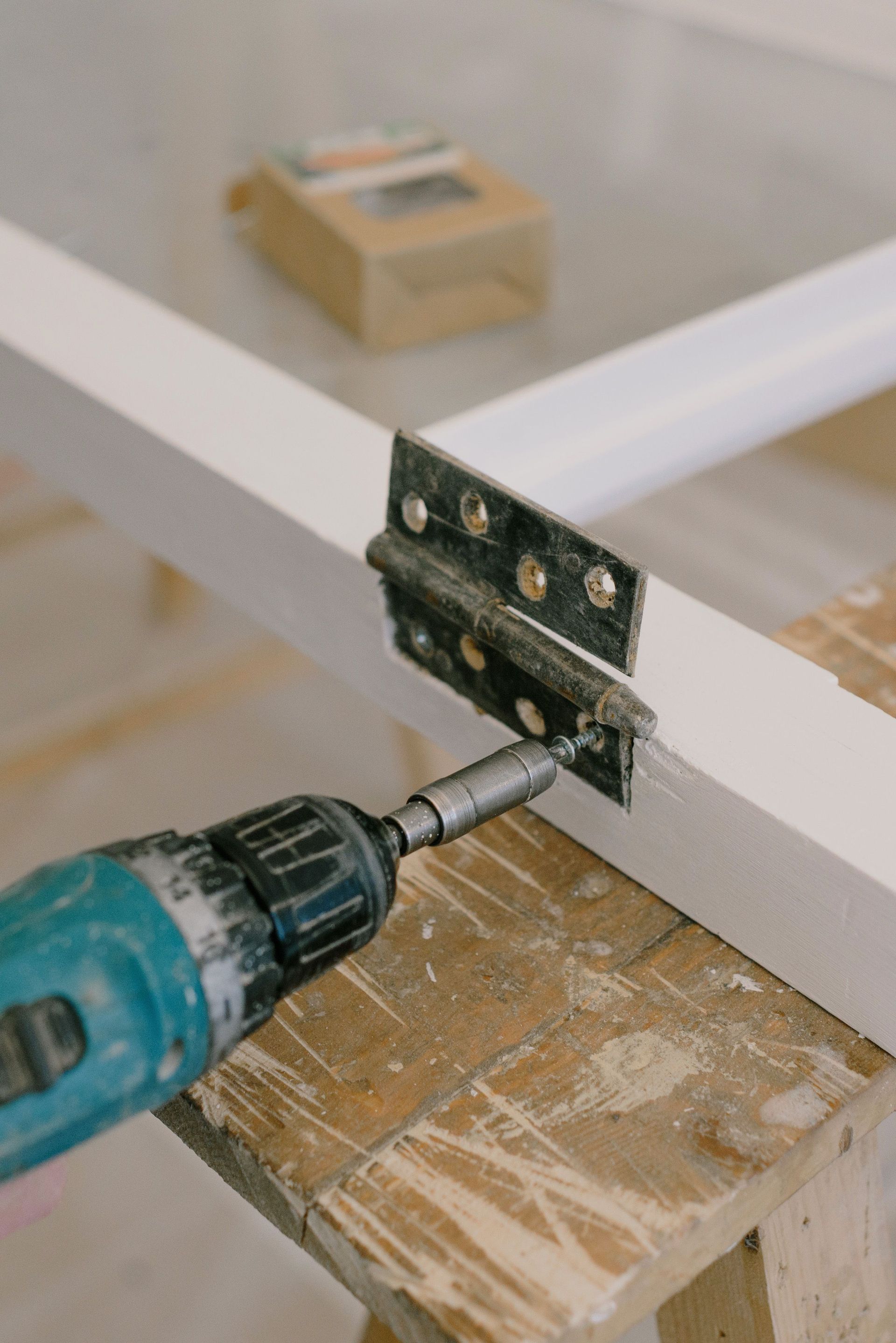 Electric screwdriver with drill attached to metal hinge on painted window placed on wooden workbench in room during relocation works